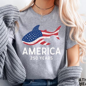 Puede incluir: Camiseta gris jaspeado con un dise&ntilde;o de tibur&oacute;n en rojo, blanco y azul, que representa la bandera americana. La palabra "AMERICA" est&aacute; impresa debajo, junto con "250 YEARS". Una prenda casual para el d&iacute;a a d&iacute;a.