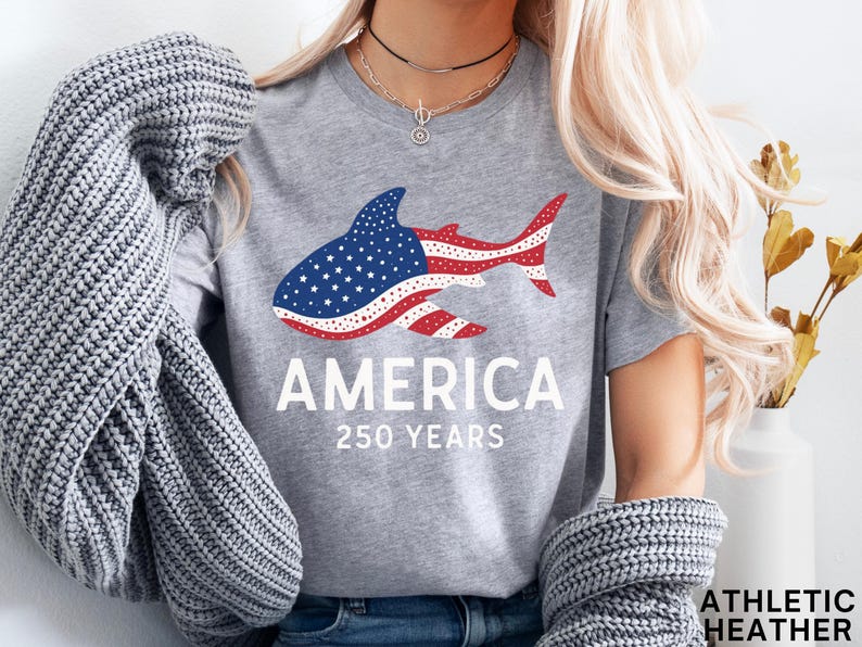Puede incluir: Camiseta gris jaspeado con un dise&ntilde;o de tibur&oacute;n en rojo, blanco y azul, que representa la bandera americana. La palabra "AMERICA" est&aacute; impresa debajo, junto con "250 YEARS". Una prenda casual para el d&iacute;a a d&iacute;a.