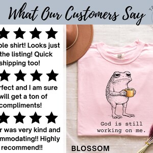 May include: A light pink t-shirt with a graphic of a grumpy frog holding a coffee cup. The text "God is still working on me" is below the frog. The word "BLOSSOM" is at the bottom of the shirt.