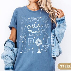 May include: A steel blue t-shirt with a white graphic design that says "Catholic Mama" with the names Amelia and John underneath. The graphic also includes a dove, a cross, a rosary, a heart, a seashell, and a bow.