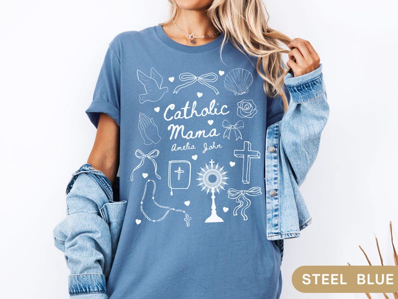 May include: A steel blue t-shirt with a white graphic design that says "Catholic Mama" with the names Amelia and John underneath. The graphic also includes a dove, a cross, a rosary, a heart, a seashell, and a bow.