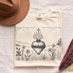 K&ouml;nnte beinhalten: Elfenbeinfarbenes T-Shirt mit einer detaillierten Schwarz-Wei&szlig;-Illustration eines Herzens mit Flammen, einem Kreuz und einer Dornenkrone. Das Design ist vor einer Landschaft mit Blumen und Himmel platziert.