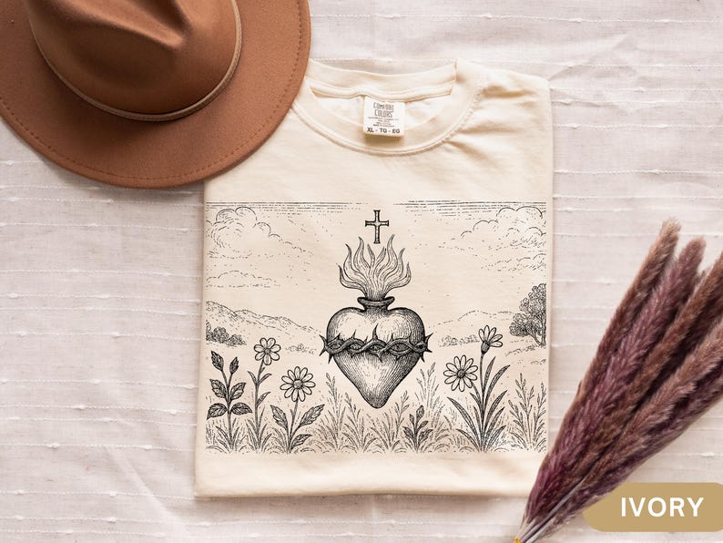 K&ouml;nnte beinhalten: Elfenbeinfarbenes T-Shirt mit einer detaillierten Schwarz-Wei&szlig;-Illustration eines Herzens mit Flammen, einem Kreuz und einer Dornenkrone. Das Design ist vor einer Landschaft mit Blumen und Himmel platziert.