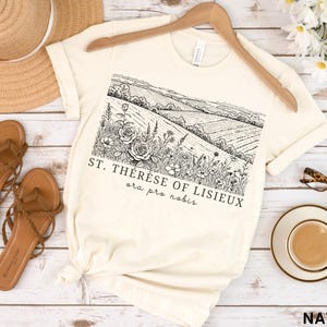 Pode incluir: Camiseta creme com uma ilustra&ccedil;&atilde;o de paisagem em preto e branco e o texto "ST. TH&Eacute;R&Egrave;SE OF LISIEUX ora pro nobis." A camisa &eacute; exibida com sand&aacute;lias, um chap&eacute;u de palha e &oacute;culos.