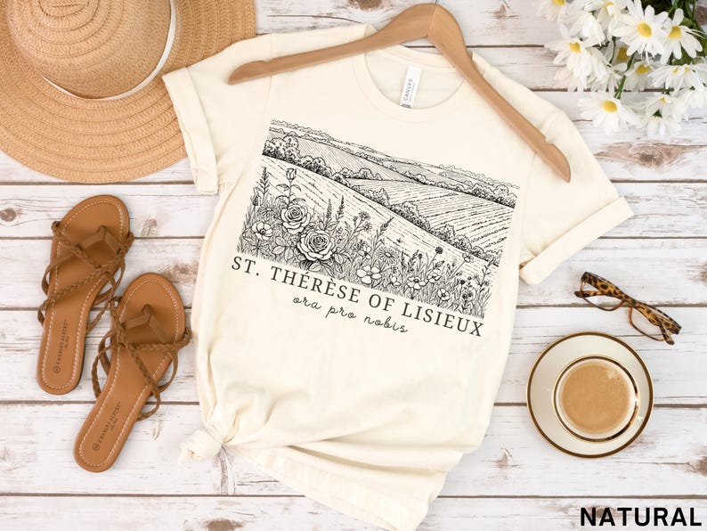 Pode incluir: Camiseta creme com uma ilustra&ccedil;&atilde;o de paisagem em preto e branco e o texto "ST. TH&Eacute;R&Egrave;SE OF LISIEUX ora pro nobis." A camisa &eacute; exibida com sand&aacute;lias, um chap&eacute;u de palha e &oacute;culos.