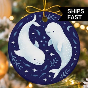 May include: A round ceramic ornament featuring a dark blue background with two white beluga whales, small fish, and star shapes. The ornament is suspended by a gold string and includes a black banner with the white text SHIPS FAST.