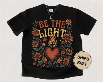 Könnte beinhalten: Schwarzes T-Shirt mit dem Text "BE THE LIGHT" in Orange und Gelb, umgeben von floralen Illustrationen. Ein Herz mit Flammen befindet sich in der Mitte. Eine silberne Halskette mit Sternanhänger ist sichtbar.