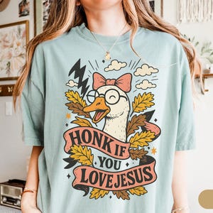 Puede incluir: Camiseta azul claro con un dibujo de un ganso de dibujos animados con gafas y un lazo rosa. El ganso est&aacute; rodeado de hojas y rayos, con el texto "HONK IF YOU LOVE JESUS" en una pancarta.