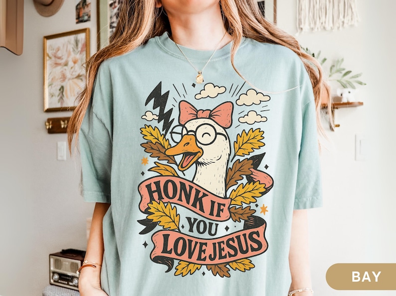 Puede incluir: Camiseta azul claro con un dibujo de un ganso de dibujos animados con gafas y un lazo rosa. El ganso est&aacute; rodeado de hojas y rayos, con el texto "HONK IF YOU LOVE JESUS" en una pancarta.
