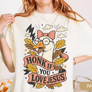 Puede incluir: Camiseta color marfil con un dibujo de un ganso de dibujos animados con gafas y un lazo, con el texto "HONK IF YOU LOVE JESUS" en una pancarta. El dise&ntilde;o incluye rayos, nubes y hojas de oto&ntilde;o.