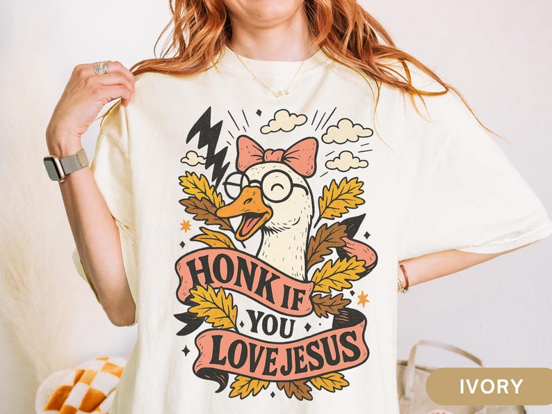 Puede incluir: Camiseta color marfil con un dibujo de un ganso de dibujos animados con gafas y un lazo, con el texto "HONK IF YOU LOVE JESUS" en una pancarta. El dise&ntilde;o incluye rayos, nubes y hojas de oto&ntilde;o.