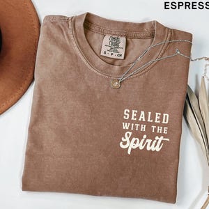 May include: Espresso-colored t-shirt with the text "SEALED WITH THE Spirit" in white. A silver necklace with a pendant is draped across the shirt. A brown hat is visible in the upper left corner.
