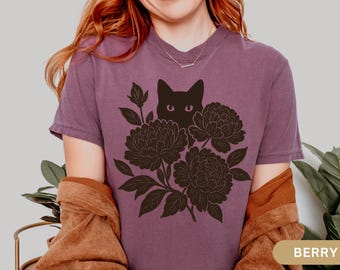 Può includere: T-shirt color bacca con un disegno di un gatto nero che sbuca da dietro dei fiori. Il motivo floreale è dettagliato con foglie. La maglietta ha un girocollo e maniche corte.