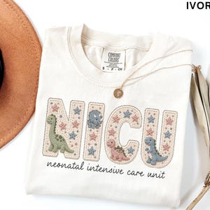 May include: An ivory t-shirt featuring the text NICU in large, block-style letters filled with a stitched pattern of small stars and dinosaurs in muted green, pink, and blue. Below the letters, the text reads neonatal intensive care unit.