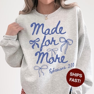 Puede incluir: Sudadera gris claro con la frase azul "Made for More" con ilustraciones de lazos. El texto "Ephesians 2:10" est&aacute; debajo. Un c&iacute;rculo rojo con "SHIPS FAST!" est&aacute; a la derecha de la sudadera.