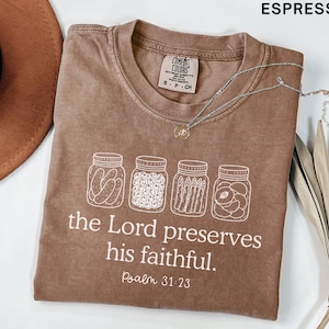 Puede incluir: Una camiseta color espresso con un gr&aacute;fico blanco de cuatro frascos de vidrio llenos de pepinillos, bayas, esp&aacute;rragos y rodajas de fruta. Debajo de los frascos, el texto dice: "the Lord preserves his faithful. Psalm 31:23." Tiene cuello redondo.