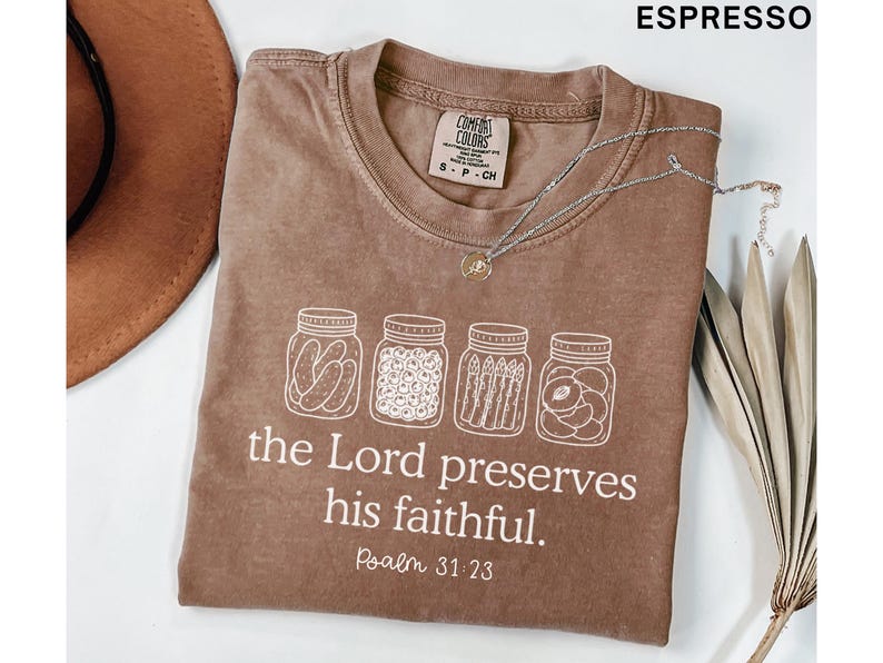Puede incluir: Una camiseta color espresso con un gr&aacute;fico blanco de cuatro frascos de vidrio llenos de pepinillos, bayas, esp&aacute;rragos y rodajas de fruta. Debajo de los frascos, el texto dice: "the Lord preserves his faithful. Psalm 31:23." Tiene cuello redondo.