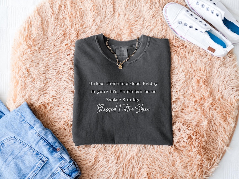 May include: Charcoal grey t-shirt featuring the quote "Unless there is a Good Friday in your life, there can be no Easter Sunday. Blessed Fulton Sheen." A gold necklace is on the shirt. White trainers and blue jeans are also visible.