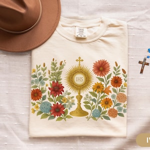 Puede incluir: Camiseta de color marfil con un diseño floral que rodea una custodia dorada con las letras IHS. También se ven un sombrero marrón y un rosario azul con una cruz.