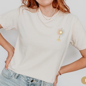 Peut inclure: T-shirt ivoire avec un motif color&eacute; et d&eacute;taill&eacute; d'un symbole religieux sur le haut de la poitrine gauche. Le motif comprend un motif de soleil aux couleurs pastel et une base dor&eacute;e. Le t-shirt est de style d&eacute;contract&eacute; &agrave; manches courtes.