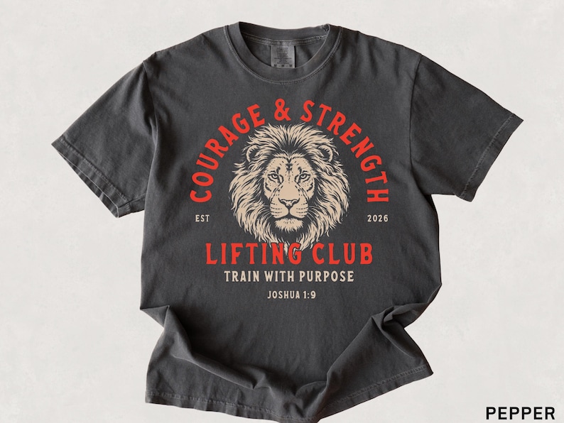 May include: Pepper-colored t-shirt with a distressed graphic. The design features a lion's head within a red circle, with the words "COURAGE & STRENGTH" and "LIFTING CLUB". The text "TRAIN WITH PURPOSE" and "Joshua 1:9" are also included.