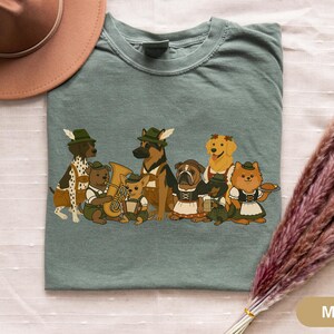 May include: Moss green t-shirt featuring a cartoon illustration of dogs dressed in traditional Bavarian outfits. The dogs include a Dalmatian, German Shepherd, Bulldog, and Golden Retriever. Some dogs are playing instruments.