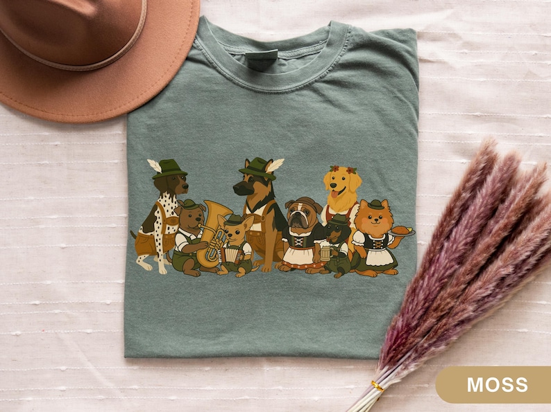 May include: Moss green t-shirt featuring a cartoon illustration of dogs dressed in traditional Bavarian outfits. The dogs include a Dalmatian, German Shepherd, Bulldog, and Golden Retriever. Some dogs are playing instruments.