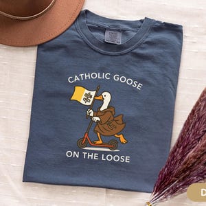 May include: Denim blue t-shirt with the text "Catholic Goose On The Loose". The graphic features a goose in a brown robe riding a scooter and holding a flag. A brown hat and dried flowers are in the background.