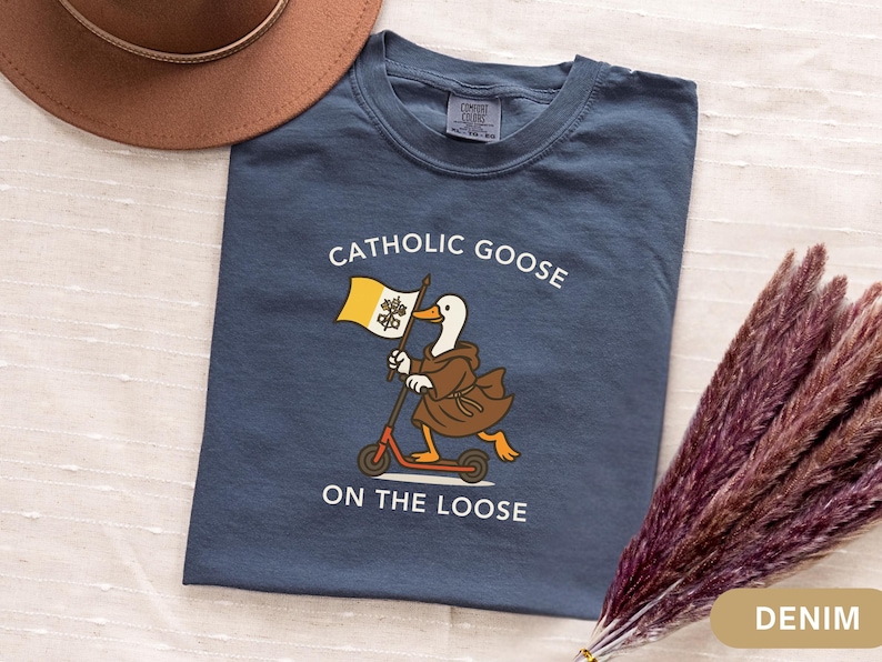 May include: Denim blue t-shirt with the text "Catholic Goose On The Loose". The graphic features a goose in a brown robe riding a scooter and holding a flag. A brown hat and dried flowers are in the background.
