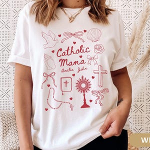 May include: White t-shirt with a red design featuring the text "Catholic Mama" with the names Amelia and John, and various religious symbols including a dove, a cross, a rosary, a bible, a heart, a seashell, a flower, and a bow.