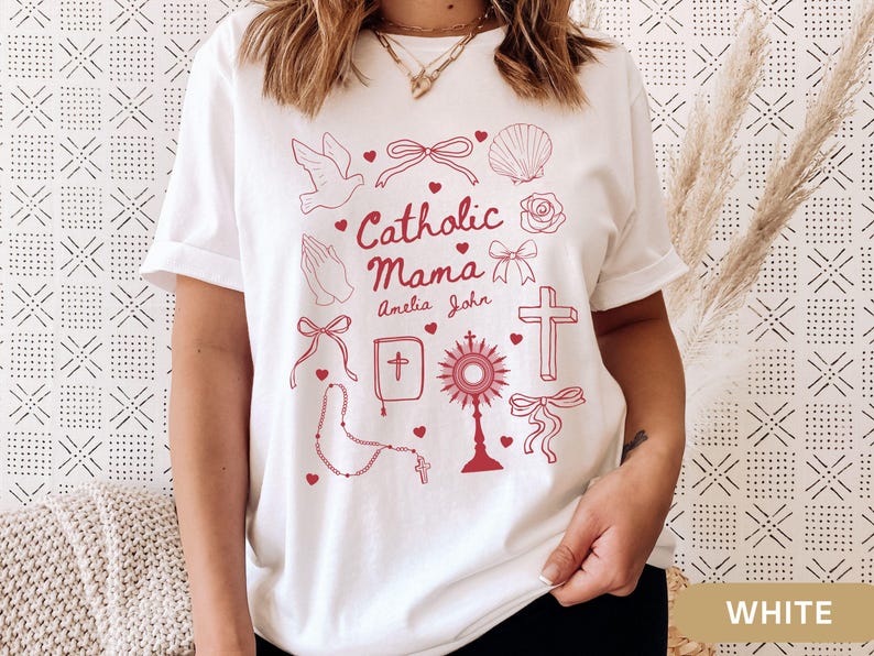 May include: White t-shirt with a red design featuring the text "Catholic Mama" with the names Amelia and John, and various religious symbols including a dove, a cross, a rosary, a bible, a heart, a seashell, a flower, and a bow.