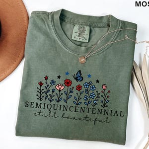 May include: Moss green t-shirt with a floral design featuring red, pink, and blue flowers, a butterfly, and the text "SEMIQUINCENTENNIAL still beautiful." A gold necklace and a brown hat are also visible.