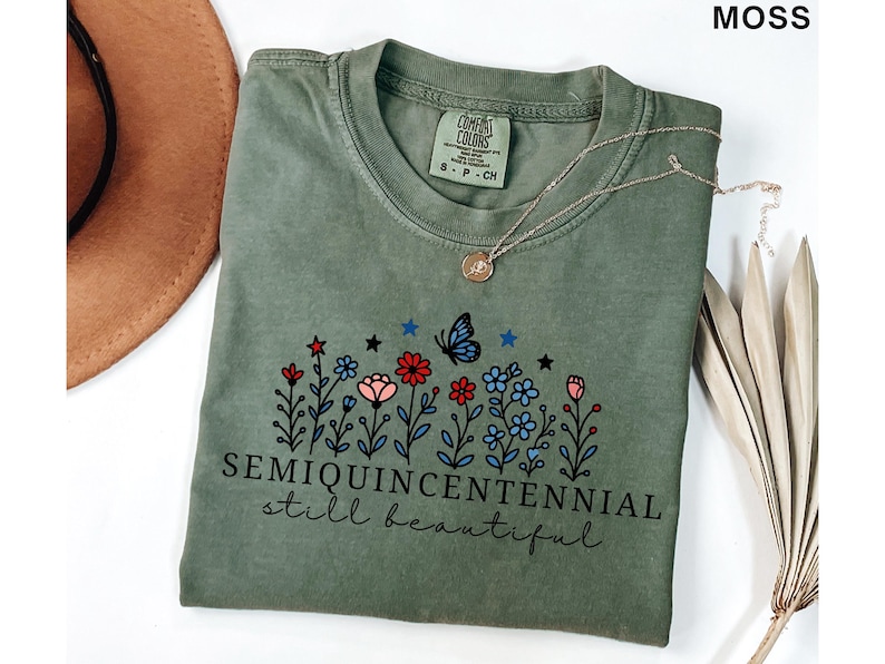 May include: Moss green t-shirt with a floral design featuring red, pink, and blue flowers, a butterfly, and the text "SEMIQUINCENTENNIAL still beautiful." A gold necklace and a brown hat are also visible.