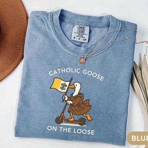 May include: Blue jean-colored t-shirt with the text "Catholic Goose On The Loose" and a cartoon goose riding a scooter while holding a flag. A gold necklace and a brown hat are also visible.