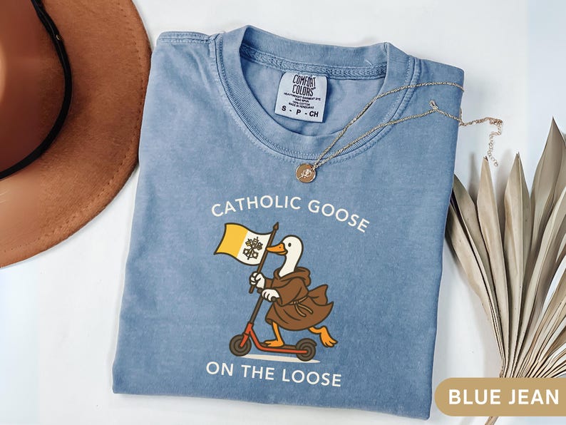 May include: Blue jean-colored t-shirt with the text "Catholic Goose On The Loose" and a cartoon goose riding a scooter while holding a flag. A gold necklace and a brown hat are also visible.