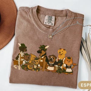 May include: Espresso-colored Comfort Colors t-shirt featuring a cartoon band of dogs in traditional German attire. The dogs are playing instruments and wearing hats. A silver necklace with a small pendant is on the shirt.