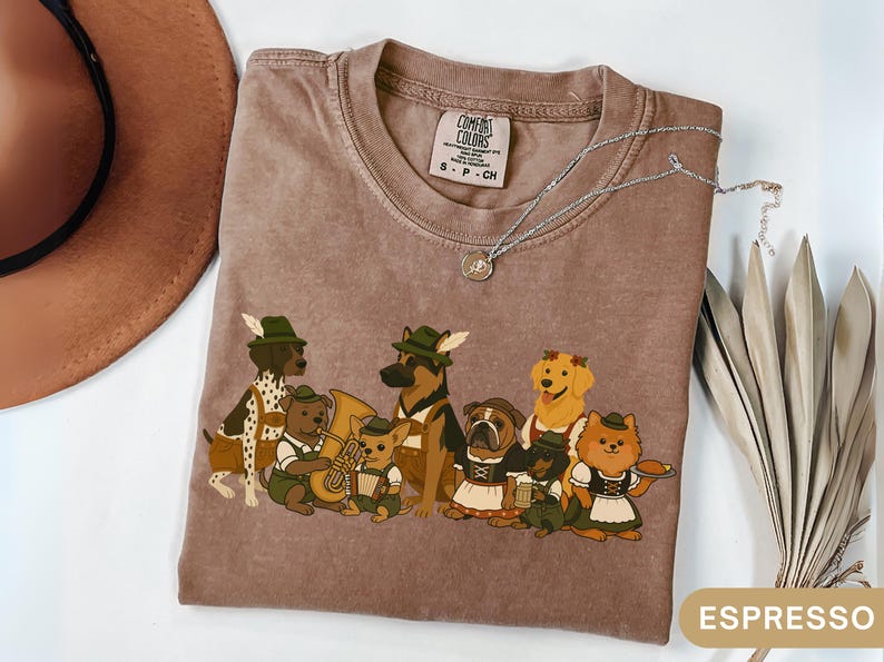 May include: Espresso-colored Comfort Colors t-shirt featuring a cartoon band of dogs in traditional German attire. The dogs are playing instruments and wearing hats. A silver necklace with a small pendant is on the shirt.