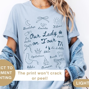Puede incluir: Camiseta azul claro con un estampado gr&aacute;fico de Nuestra Se&ntilde;ora en gira. El gr&aacute;fico incluye varios iconos y lugares religiosos, como Guadalupe, Lourdes, F&aacute;tima, Par&iacute;s, La Salette, Czestochowa, Akita, Knock y Champion.