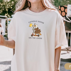 May include: Ivory-colored t-shirt featuring a cartoon goose riding a scooter and holding a flag. The text on the shirt reads "Catholic Goose On The Loose". The design is in shades of brown, yellow, and black.