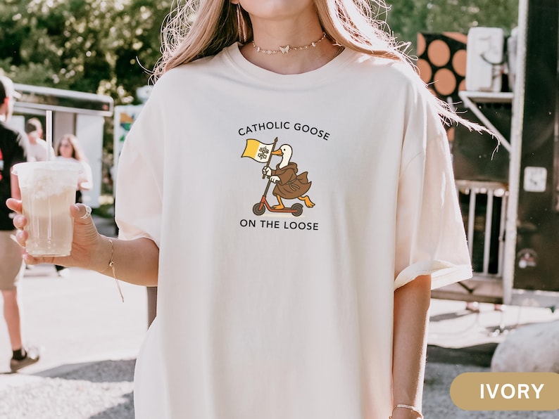 May include: Ivory-colored t-shirt featuring a cartoon goose riding a scooter and holding a flag. The text on the shirt reads "Catholic Goose On The Loose". The design is in shades of brown, yellow, and black.