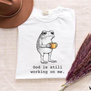 May include: White t-shirt featuring a grumpy cartoon frog holding a coffee cup. The frog is drawn in black and white, with the coffee cup in a warm yellow-orange. The text "God is still working on me" is printed below the frog.