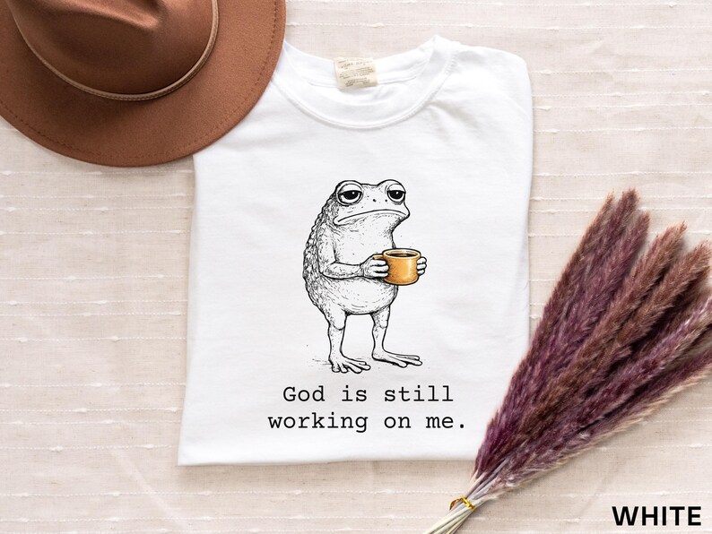 May include: White t-shirt featuring a grumpy cartoon frog holding a coffee cup. The frog is drawn in black and white, with the coffee cup in a warm yellow-orange. The text "God is still working on me" is printed below the frog.