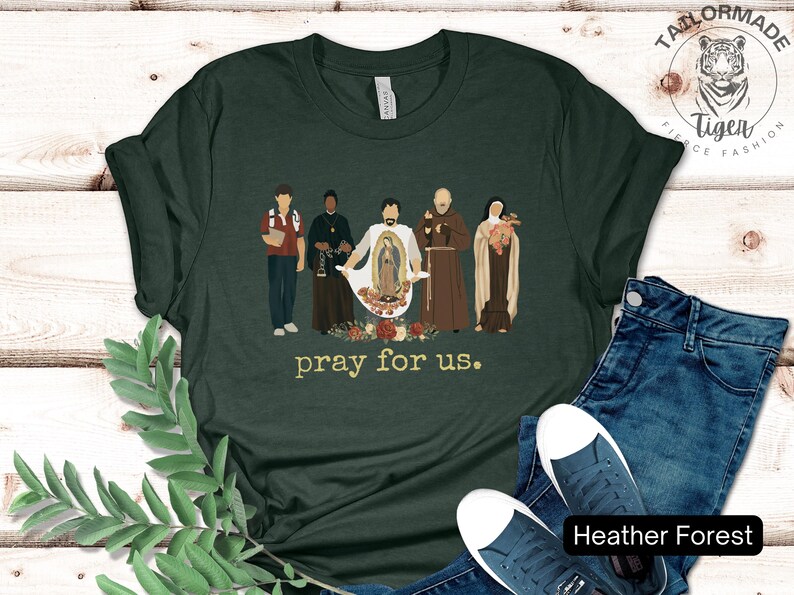 May include: A dark green t-shirt with a graphic of five figures standing in front of a floral design. The text "pray for us." is printed below the graphic.