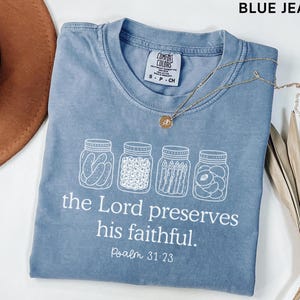 Puede incluir: Una camiseta color azul jean con un gr&aacute;fico blanco de cuatro frascos de vidrio llenos de conservas. Debajo de los frascos, el texto dice: "the Lord preserves his faithful. Psalm 31:23". La camiseta tiene una etiqueta de la marca Comfort Colors.