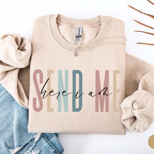 May include: A beige sweatshirt with the text "SEND ME here i am" in colorful letters. The text is in a rainbow color scheme with pink, orange, yellow, green, blue, and purple.