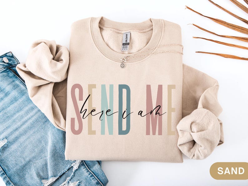 May include: A beige sweatshirt with the text "SEND ME here i am" in colorful letters. The text is in a rainbow color scheme with pink, orange, yellow, green, blue, and purple.