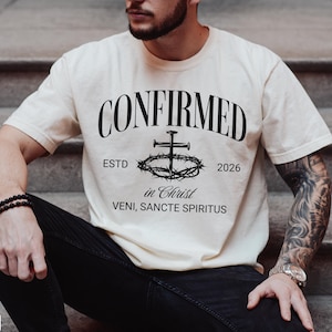 Può includere: Una t-shirt a maniche corte color avorio con testo nero che recita CONFIRMED, ESTD 2026, in Christ e VENI, SANCTE SPIRITUS. La grafica include un'illustrazione centrale di una croce fatta di chiodi sopra una corona di spine.