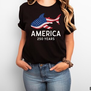 Puede incluir: Camiseta negra con un dise&ntilde;o de tibur&oacute;n con los colores de la bandera estadounidense. La palabra "AMERICA" est&aacute; impresa encima de "250 YEARS" en blanco. La modelo lleva vaqueros azules.