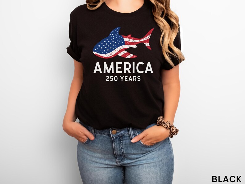 Puede incluir: Camiseta negra con un dise&ntilde;o de tibur&oacute;n con los colores de la bandera estadounidense. La palabra "AMERICA" est&aacute; impresa encima de "250 YEARS" en blanco. La modelo lleva vaqueros azules.