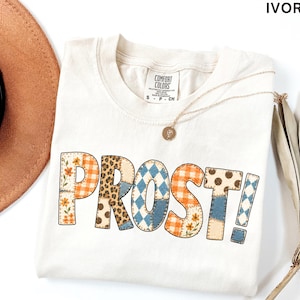 May include: An ivory short-sleeve t-shirt featuring the word PROST! in large, block-style letters. Each letter has a unique patchwork pattern, including orange gingham, leopard print, blue argyle, and floral designs. The shirt has a crew neckline.